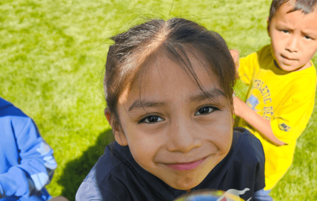 Close-up of a smiling young child looking directly at the camera on a grassy field, with two other children partially visible behind them.