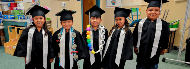 Five children wearing graduation caps and gowns stand together in a classroom, smiling for a photo.